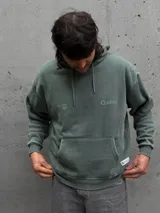 Canguro con capucha de color verde con efecto lavado, diseño de corte holgado, bolsillo frontal tipo canguro, cordones ajustables y logo estampado en el pecho.