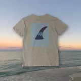 Remera gris con estampado en la espalda de una aleta de surf con un ojo en el centro, dentro de un marco celeste con motivos geométricos.