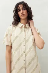 Vestido camisero color crema con rayas diagonales verdes, de corte recto, con cuello camisero, mangas cortas con dobladillo, cierre frontal con botones y dos bolsillos de parche en el pecho.