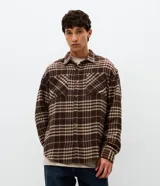 Camisa tipo overshirt de corte oversized, confeccionada en tejido de poliéster con textura. Presenta un estampado de cuadros en tonos marrones y beige, cuello clásico, cierre frontal con botones y bolsillo aplicado en el pecho.