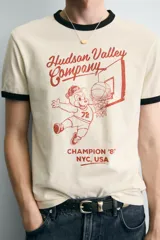 Camiseta de corte regular fit, color crema, con cuello redondo y mangas cortas. Presenta un estampado frontal con un personaje de dibujos animados jugando al baloncesto y texto "Hudson Valley Company Champion '87 NYC, USA". Los acabados del cuello y las mangas son de vivo negro a contraste.