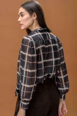 Camisa de manga larga con estampado a cuadros, cuello alto y cierre frontal con botones. Presenta un diseño fluido con lazo decorativo en el cuello y puños abotonados.