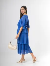 Vestido midi de seda italiana color azul, con escote en V, mangas murciélago amplias, falda de tres volados y cintura elastizada con lazo ajustable al tono y hebilla circular dorada.