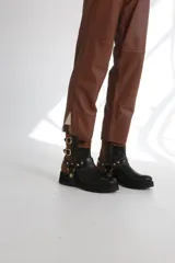 Bota estilo biker de caña media, confeccionada en cuero negro con detalles en gamuza marrón. Presenta correas ajustables con hebillas metálicas, tachas decorativas y suela track dentada.