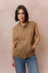 Chaleco de gamuza color marrón con acabado aterciopelado, cuello alto con broche a presión, cierre frontal metálico y bolsillos laterales con cierre.