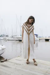 Vestido camisero largo de lino color beige, con botones frontales, mangas cortas y volados dispuestos en líneas angulares sobre el pecho y los hombros.