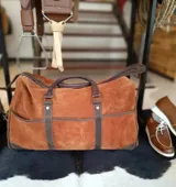 Bolso de mano grande tipo duffel, confeccionado en gamuza marrón con detalles de cuero oscuro en las asas, base y correas verticales de refuerzo. Cuenta con amplios bolsillos frontales abiertos y cierre superior con cremallera.