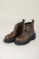 Bota corta de cuero con efecto gastado, diseño de puntera cuadrada y suela track robusta de goma negra. Presenta una hebilla metálica decorativa en el empeine y tirador en la parte trasera.