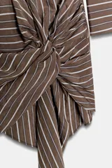 Vestido corto de estilo camisero con estampado de rayas verticales, cuello solapa, manga larga y cierre frontal con botones. Presenta un detalle de lazada en la cintura que crea un efecto drapeado y un bajo asimétrico.