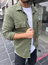Sobre camisa verde militar con cuello clásico, cierre frontal con botones, dos bolsillos con solapa abotonada en el pecho y mangas largas con puños abotonados.