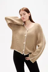 Cardigan de punto color natural, con cuello en V, cierre frontal con botones y diseño holgado. Presenta detalles de costuras decorativas bordadas a lo largo de las mangas y en los bordes.