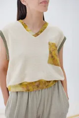 Chaleco de lana merino color crema, con escote en V y diseño sin mangas. Se usa sobre una camisa mostaza de mangas largas con puños elásticos.