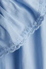 Vestido corto de algodón celeste, con cuello alto con volados y abertura frontal con botones. Presenta mangas largas, elástico en la cintura y falda escalonada con detalles de encaje y volados en los bordes.