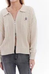 Cardigan de tejido de punto color marrón, con cuello solapa, cierre frontal de cremallera y diseño de trenzas. Cuenta con puños y dobladillo en rib elástico y un pequeño bordado en el pecho.