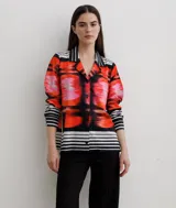 Camisa de manga larga con cuello solapa y cierre frontal de botones. Presenta un diseño estampado con motivos abstractos en tonos rojos, naranjas y rosas, combinado con detalles de rayas horizontales en blanco y negro en el cuello, puños y ruedo.