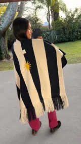 Ruana de punto a rayas verticales negras y beige, confeccionada en hilo de algodón, con bordado artesanal de sol en la espalda y flecos en el ruedo.
