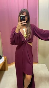 Vestido largo color bordó con escote en V profundo, detalle de nudo central y aberturas laterales en la cintura. Presenta mangas largas abullonadas con puños elásticos y falda con tajo frontal pronunciado.