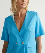 Blazer celeste de lino con escote en V, manga corta, cierre frontal con dos botones dorados y bolsillos de solapa.