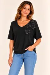 Remera negra de punto liviano con escote en V y mangas cortas. Presenta aplicaciones de flores tridimensionales y pequeñas cuentas decorativas en el hombro y pecho izquierdo.