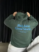 Canguro verde oliva con capucha y cordones ajustables. Estampado en la espalda con la frase "Bad Ideas Good Stories" en letras azules.