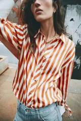 Camisa de manga larga con estampado de rayas verticales en color naranja y crema. Presenta cuello solapa, cierre frontal con botones dorados y puños anchos en color crema con detalle de lazos ajustables.
