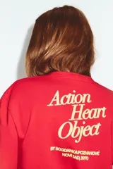 Camiseta de algodón color rojo, corte regular fit, cuello redondo y manga corta. Presenta una estampa fotográfica en el centro con dos personas sosteniendo un gran corazón rojo inflable. Incluye texto con nombre y copyright.