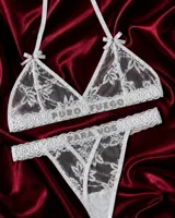 Conjunto de lencería de tul bordado blanco, compuesto por un corpiño triangular con tirantes halter, moños decorativos y la inscripción 'PURO FUEGO' en strass, y una bombacha tipo colaless con tiras cruzadas en el frente y la inscripción 'PARA VOS' en strass.