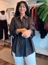 Camisa blanca de corte clásico y diseño holgado, con cuello camisero que presenta un detalle de pedrería o brillo sutil en los bordes. Posee mangas largas con puños rectos y un largo asimétrico, siendo más corta en la parte delantera y más larga en la parte trasera.