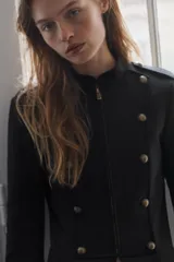 Blazer de estilo militar color negro, con cuello redondo, cierre frontal con cremallera y detalle de botones dorados decorativos en el frente. Posee mangas largas y forro interior a tono.