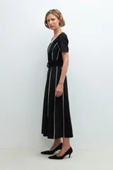 Vestido midi negro con rayas verticales blancas, escote redondo y cinturón negro con hebilla circular.