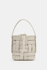 Bolso tipo bucket con cuerpo trenzado en color beige. Cuenta con asa de mano fija, correa bandolera extraíble y compartimento interior de tela con cierre fruncido.