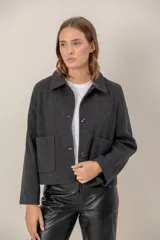 Chaqueta de paño color gris oscuro, con cuello camisero, cierre frontal de botones y dos bolsillos frontales tipo parche. Presenta un corte recto y mangas largas.