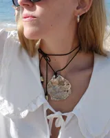 Collar artesanal con dije central de metal con textura orgánica irregular que evoca una ostra. El dije está montado sobre un cordón de algodón natural, permitiendo un uso versátil y regulable.