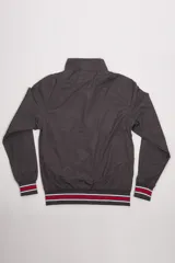 Campera liviana de nylon color gris oscuro, con cierre frontal completo, cuello subido y puños y cintura acanalados con rayas en rojo y blanco. Presenta logo bordado en el pecho y bolsillos laterales con cierre.