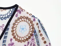 Remera corta de poliéster color crudo con bordados en tonos azules, marrones y morados, escote en V con ribete oscuro y cierre frontal con cordones y cuentas.