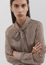 Blusa de rayas verticales marrones y blancas, con cuello alto y bufanda integrada para anudar al cuello, cierre frontal con botones y manga larga.