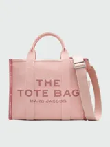 Bolso tipo tote de lona color negro con letras blancas bordadas en el frente que dicen 'THE TOTE BAG MARC JACOBS'. Cuenta con asas superiores cortas y una correa de hombro ajustable y extraíble.
