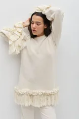 Sweater de lana merino color crudo, de silueta amplia y relajada. Presenta mangas largas con volumen, decoradas con volados y flecos en los puños. El ruedo también cuenta con detalles de volados y flecos, aportando textura y movimiento a la prenda.