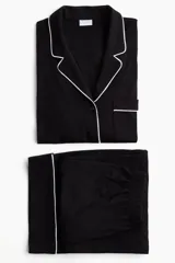 Pijama de dos piezas negro, compuesto por una camisa de manga larga con solapas de muesca, ribetes blancos y cierre de botones, y un pantalón de pierna ancha con elástico revestido en la cintura y ribetes blancos en los puños.