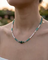 Collar de acero con baño de oro, compuesto por una combinación de perlas cultivadas blancas y piedras naturales en tonos verdes y turquesas. Presenta un dije central de piedra verde oscura y un pequeño dije colgante con logo grabado.