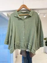 Camisa de lino color verde seco, de corte holgado y diseño corto, con cuello clásico, cierre frontal con botones y mangas tres cuartos con detalles de pliegues en los puños y el ruedo.