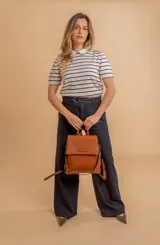 Mochila tipo cartera con tapa, confeccionada en símil cuero color suela. Cuenta con doble compartimento, división interna, cierre de seguridad y bolsillo trasero con cierre. Incluye una correa frontal decorativa con tachas metálicas.