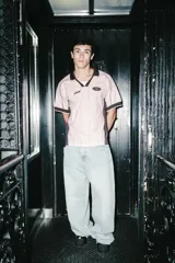 Camiseta de estilo deportivo con cuello polo, confeccionada en tela técnica de secado rápido. Presenta un diseño a rayas verticales en color rosa con paneles laterales en malla negra, mangas cortas con bordes negros y detalles gráficos estampados en el frente.