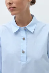 Camisa de popelina celeste con finas rayas blancas verticales, cuello camisero y abertura frontal con botones joya. Manga larga con puño ancho.