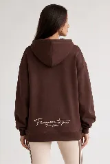 Hoodie Trena de frisa color marrón chocolate, con moldería oversize, hombros caídos, capucha con cordones ajustables y bolsillo tipo canguro. Presenta puños y ruedo de rib elástico, y detalles de bordado en la manga.