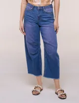 Jean wide leg cropped de tiro medio-alto, confeccionado en denim azul intermedio con efecto degradé y costuras en tono camel. Presenta cinco bolsillos, cierre frontal con botón y cierre, nervaduras decorativas en la zona de las rodillas y ruedo desflecado.