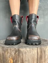 Bota de cuero con efecto desgastado en tonos marrones y negros, con suela track robusta y cierre lateral. Presenta detalles en color rojo en la parte superior de la caña.
