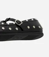 Zapato tipo salomé de color negro con acabado liso, puntera redondeada y suela plana. Presenta una tira central con hebilla metálica ajustable y está decorado con tachuelas metálicas plateadas distribuidas por toda la superficie.