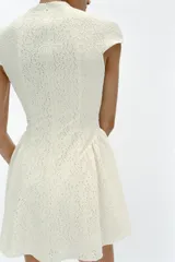 Vestido corto blanco de encaje, con cuello redondo, manga corta y bajo evasé con frunces. Cuenta con fila de falsos botones negros delanteros a contraste y cierre trasero con cremallera oculta.