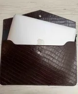 Funda para laptop de diseño minimalista, confeccionada en cuero negro con textura grabada tipo reptil y cierre mediante botón metálico a presión.